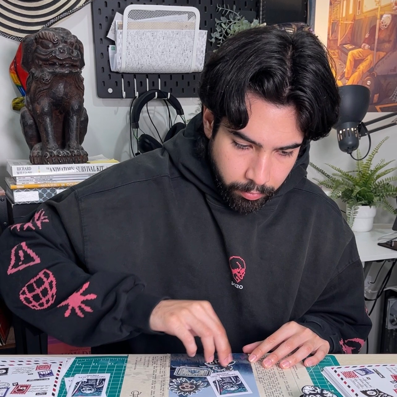 Ein Mann im schwarzen Hoodie sitzt in seinem Atelier und legt Aufkleber auf dem Tisch vor sich aus, im Hintergrund Skulpturen und Kunstwerke.