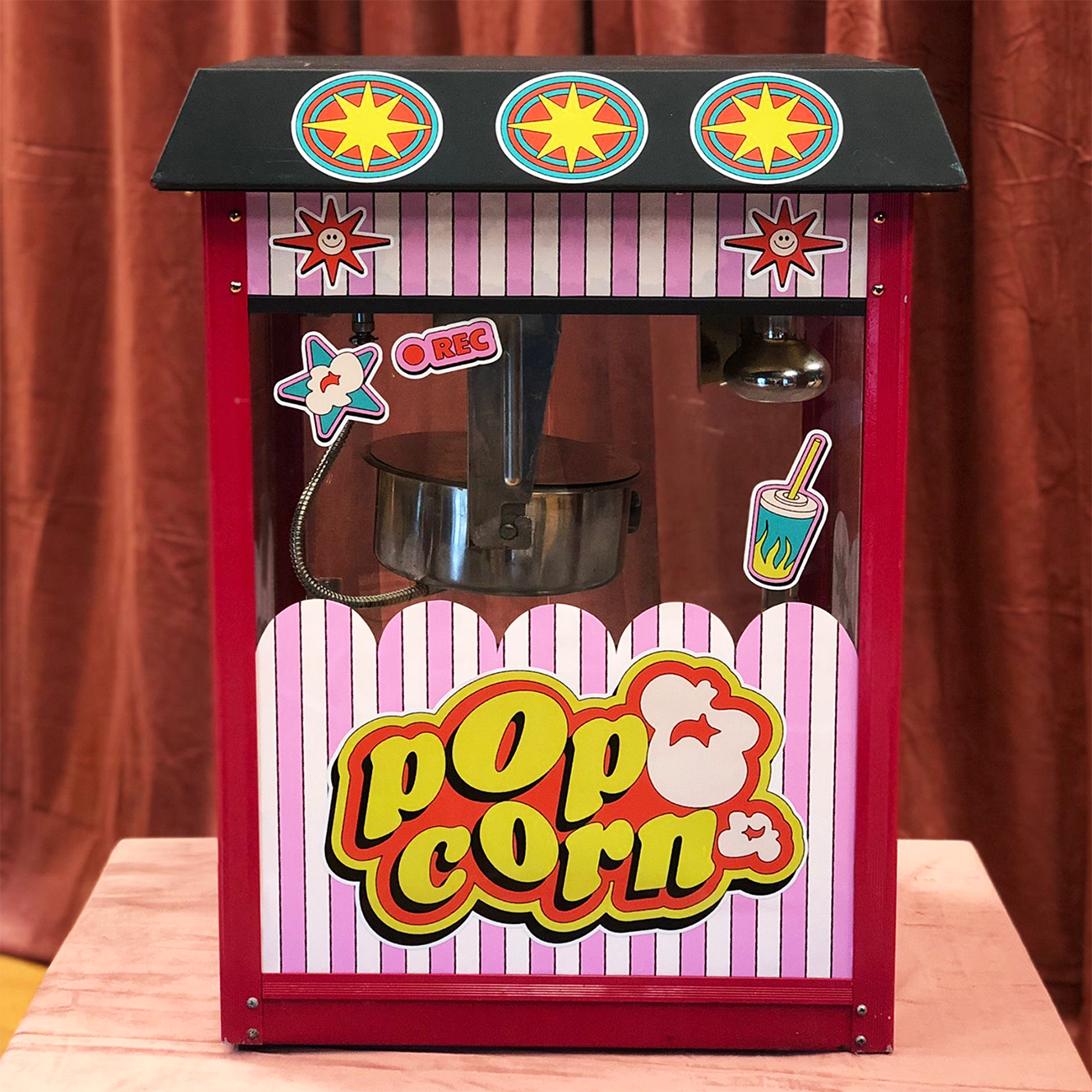 Red and pink striped popcorn machine with colorful die-cut vinyl stickers, set against a brown curtain background.