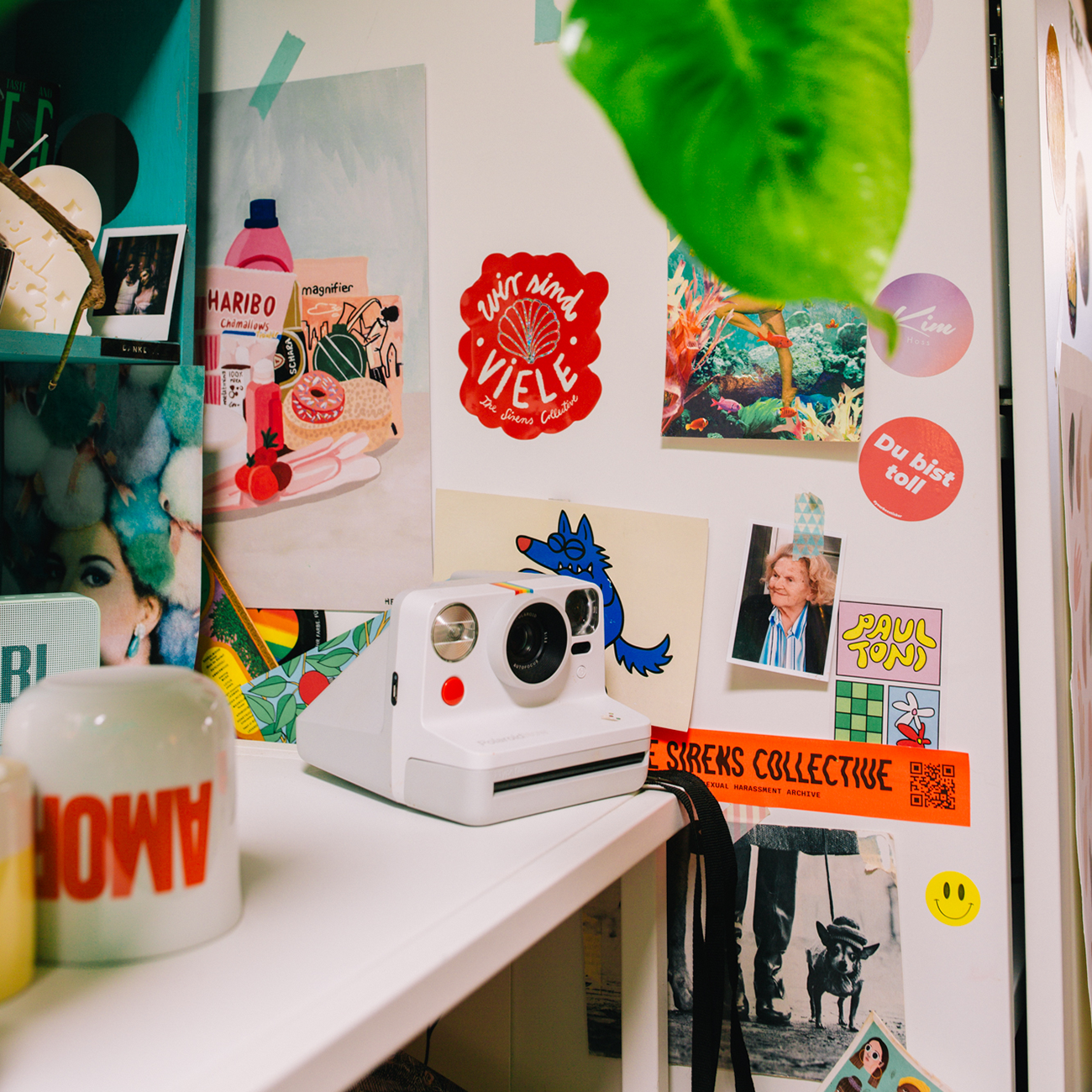 A white instant camera on a cluttered desk with colorful die-cut stickers, photos, and a plant in the background.