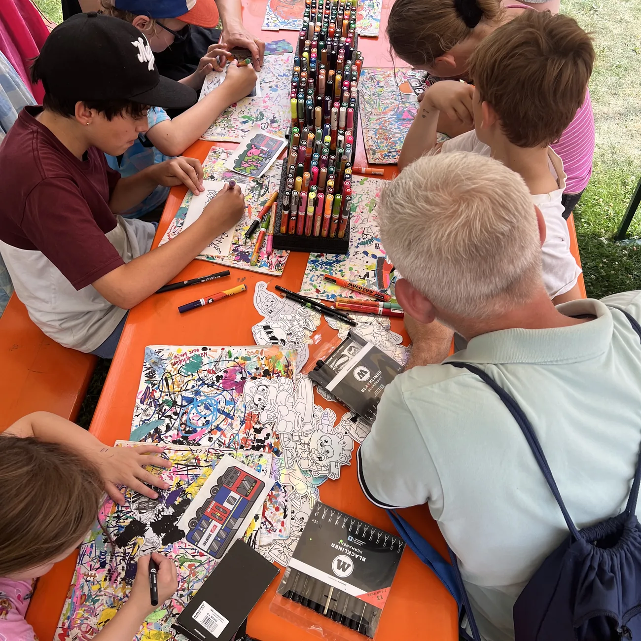 Kinder malen Freiform-Aufkleber von Brainfart rund um einen Ständer mit Paint-Markern bei einem Festival-Workshop im Freien aus