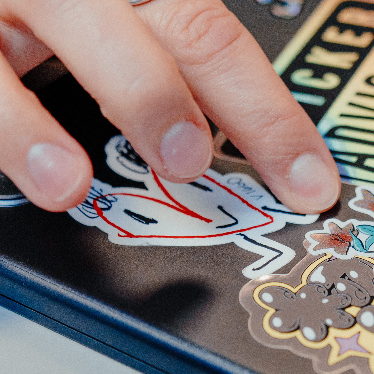 Close-up of a hand applying a white sticker with a red and black illustration onto a laptop covered with other stickers.