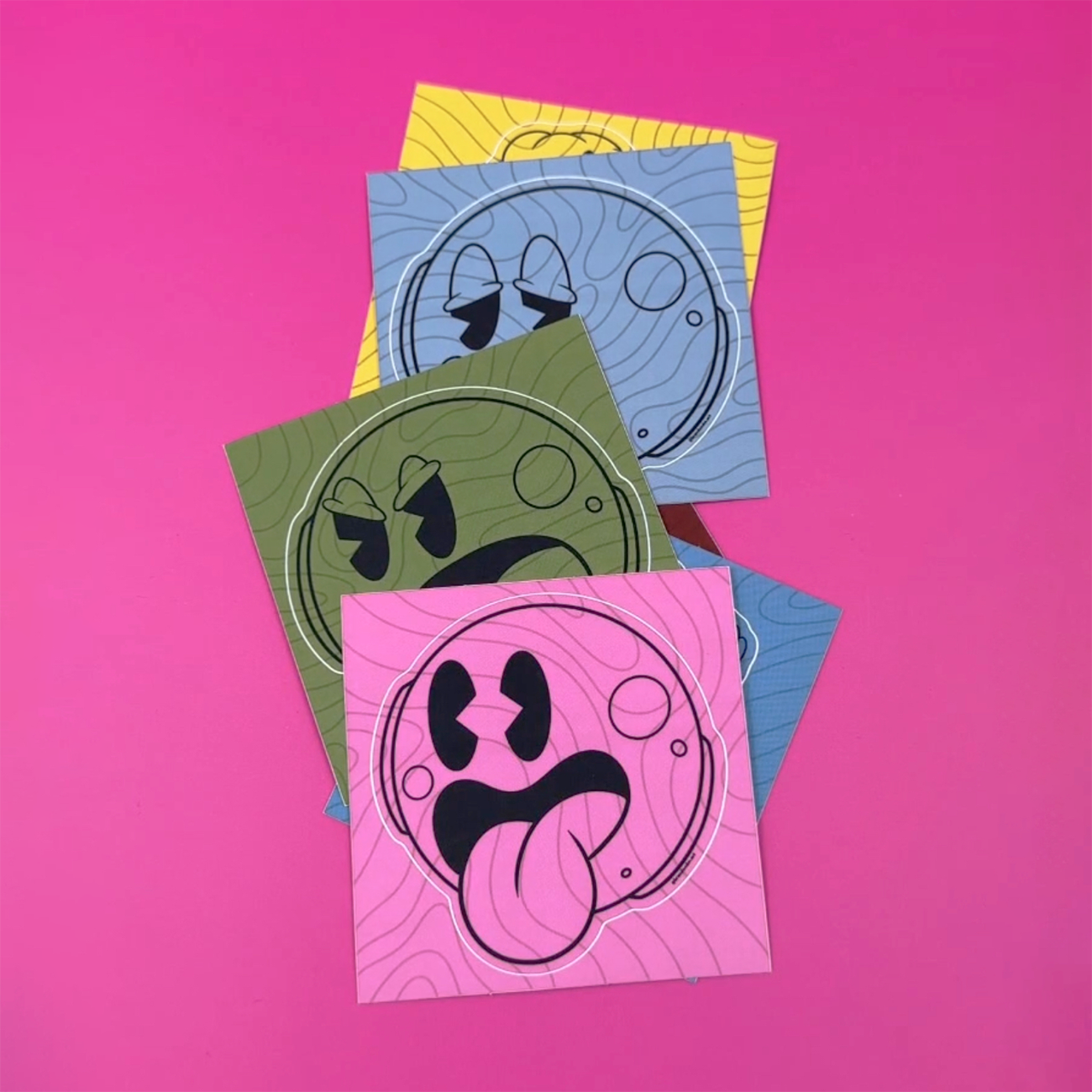 Kiss-cut sticker smiley faces in different colors and facial expressions printed on matte vinyl, against a pink background.