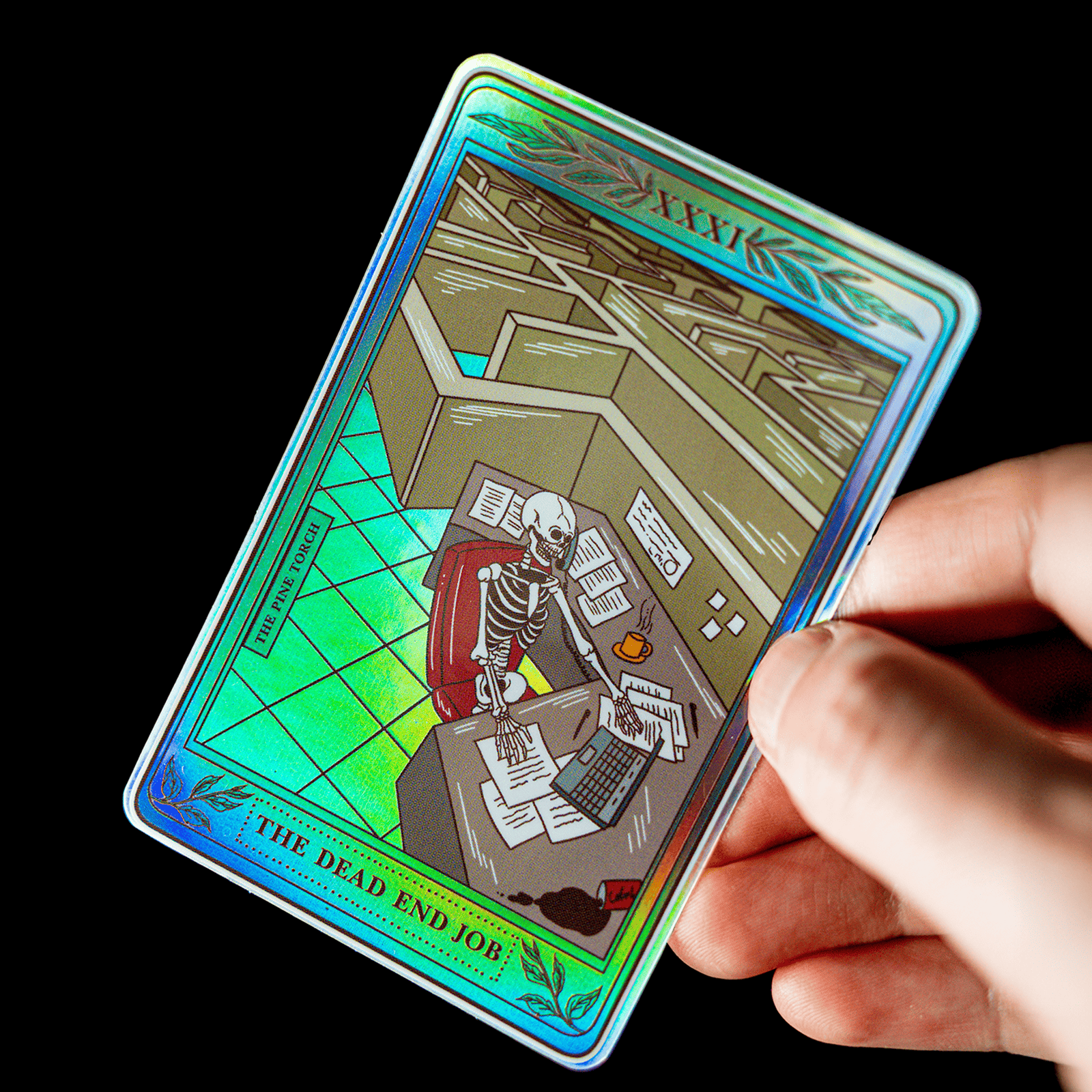 Hand hält einen holografischen Tarotkarten-Aufkleber mit dem Titel „The Dead End Job“ und einem Skelett am Bürotisch