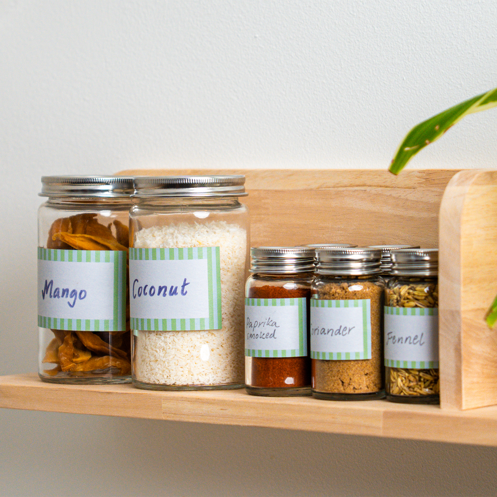 Gläser mit handbeschrifteten Etiketten auf einem Küchenregal aus Holz. Darin getrocknete Mango, Kokos und verschiedene Gewürze wie geräuchertes Paprikapulver, Koriander und Fenchelsamen.