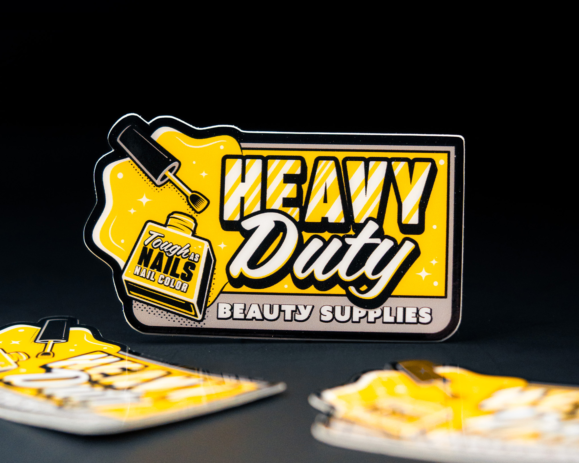 Yellow and black "Heavy Duty Beauty Supplies" sticker with a nail polish illustration and smaller sticker in foreground on black background.