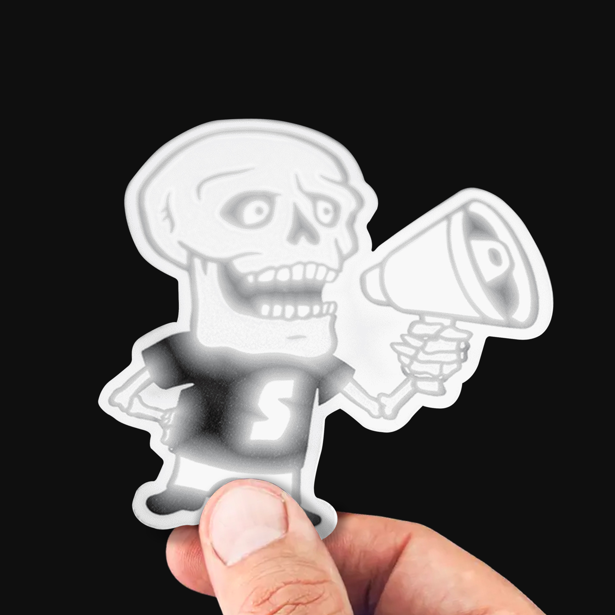Hand hält einen reflektierenden Freiform-Sticker eines Skeletts mit Megafon, das im direkten Licht weiß leuchtet