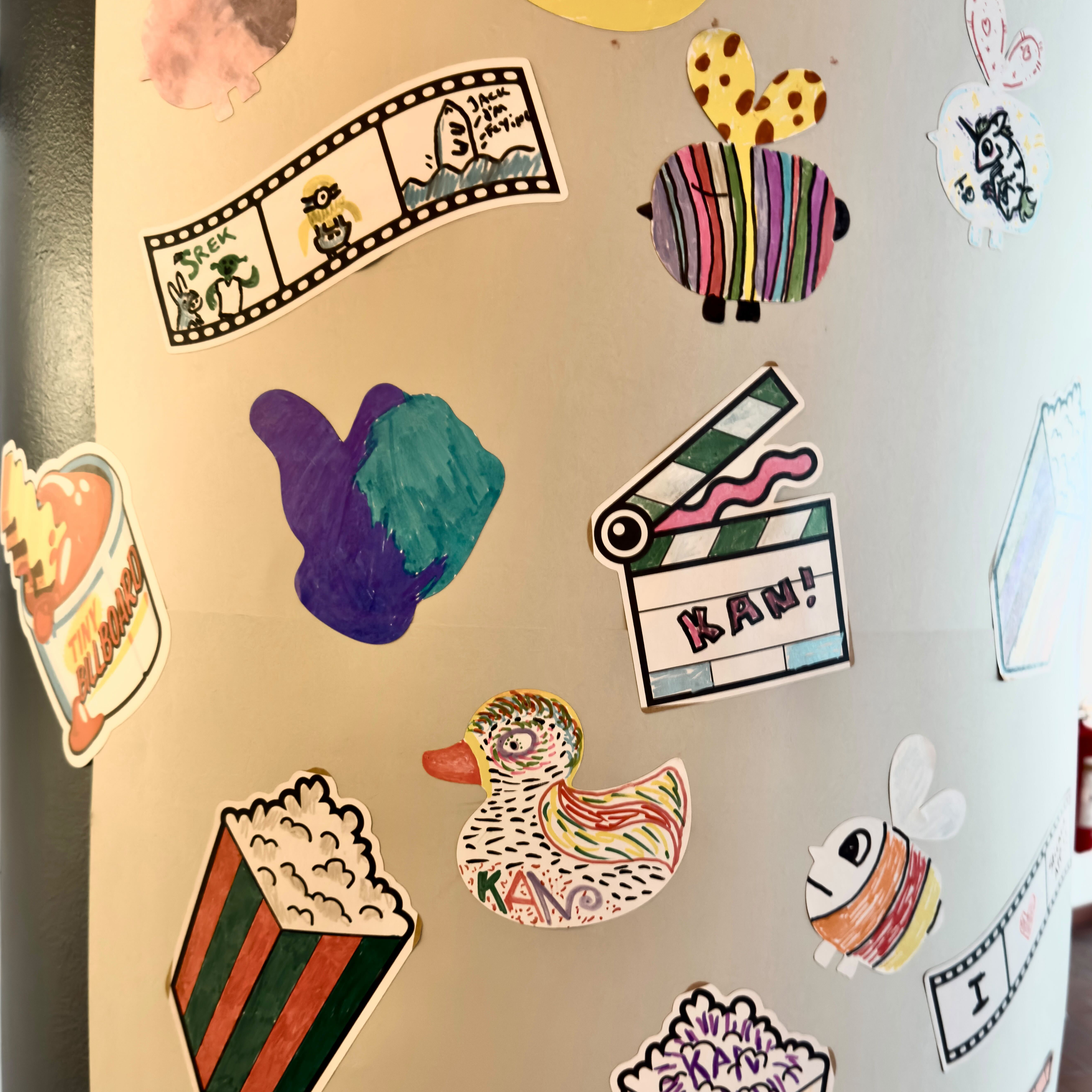Säule voller handgezeichneter Sticker, die Gäste beim KAN Film Festival gestaltet haben, darunter Popcorn, Filmstreifen, eine Ente und eine Filmklappe mit der Aufschrift „KAN!“